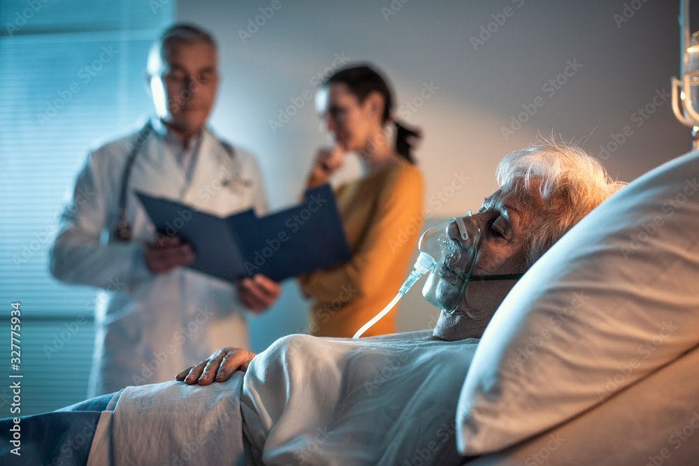 Patient in bed with ventilator with doctor and family member in background ©StockPhotoPro-stock.adobe.com